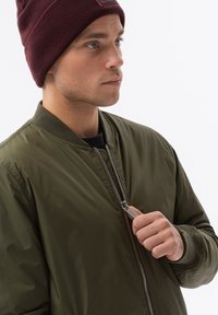Giubbotto bomber in nylon verde oliva con colletto a coste. Zip argentata in evidenza sul petto, indossato con accessori in maglia burgundy.