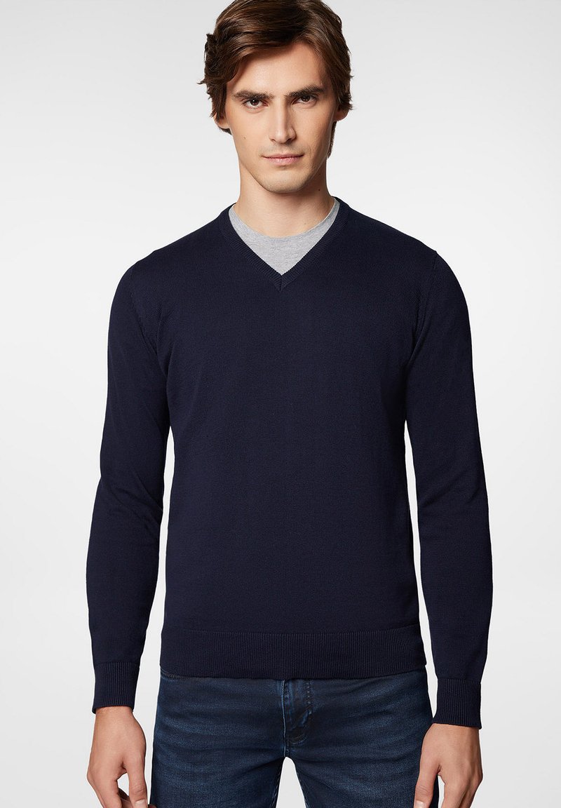 Granatowy sweter z dekoltem w serek, długimi rękawami oraz prążkowanymi mankietami i dołem. Nosi się go na szarym t-shircie, charakteryzującym się gładką, dzianinową teksturą.