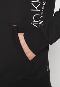 Felpa nera con tasca a marsupio, polsini a coste e logo "Calvin Klein" bianco lungo il lato. Tessuto testurizzato e morbido.