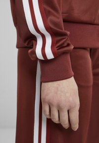 Bordeaux-coloured urheilupukukokonaisuus, jossa on valkoiset kaksoishiukset hihassa ja lahkeessa. Materiaali näyttää sileältä ja joustavalta. Näkyvä käsi hienolla tekstuurilla.
