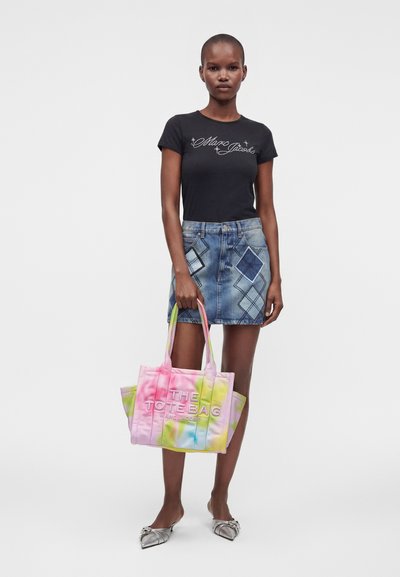 Femme portant un t-shirt noir "Marc Jacobs" et une jupe en jean, tenant un sac cabas coloré tie & dye avec des ballerines pointues argentées.