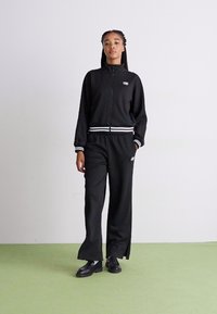 Tuta atletica nera con accenti a strisce bianche, composta da una giacca con zip e pantaloni a gamba larga. Il materiale sembra morbido; il modello indossa scarpe nere.