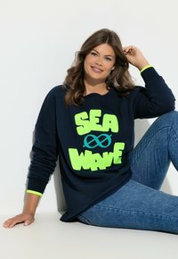 Frau mit braunen Haaren, die einen marineblauen Pullover mit neongrünem "SEA WAVE"-Schriftzug und blaue Jeans trägt, sitzt vor einer weißen Wand.