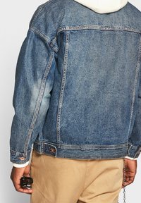 Giacca di denim blu medio con effetto sbiadito, dotata di chiusura a bottoni, design classico e polsini standard. Indossata sopra un hoodie bianco.