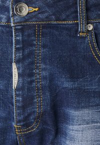 Tummansiniset farkut, joissa on haalistumismalleja. Ominaisuuksiin kuuluu keltainen saumaus ja hopeinen nappi, jossa on kuvattu brändin nimi.