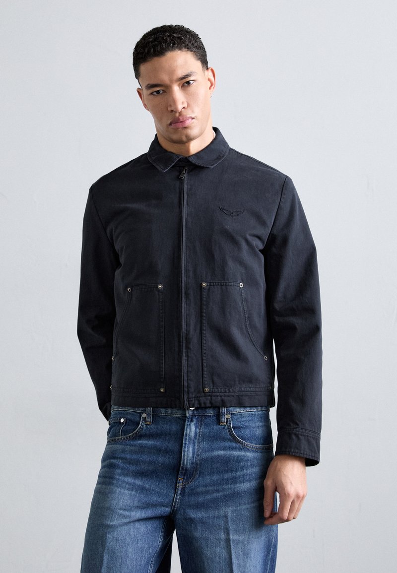 Black denim jacket with a collared neckline, front zipper, and two patch pockets. Worn over blue jeans, featuring stitching and minimalist detailing.