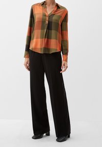Blusa a maniche lunghe, ampia e comoda, con un motivo a quadri arancione, verde e nero, dotata di colletto e pattina con tre bottoni. Abbinata a pantaloni neri.