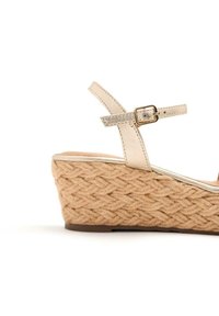Sandali con zeppa beige con una base in juta intrecciata. Presentano un cinturino metallizzato più stretto con dettaglio fibbia, in contrasto con la suola testurizzata.