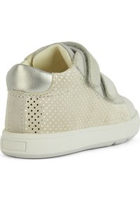 Zapatilla de ante beige con detalles perforados y acentos metálicos plateados. Presenta una puntera redondeada, correa de velcro y suela de goma blanca.