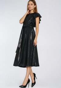 Kobieta ubrana w czarną, błyszczącą sukienkę midi z falbanami na rękawach i paskiem w talii, w zestawieniu z czarnymi butami na wysokim obcasie, stojąca na tle jednolitego tła.