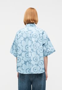 Άτομο με κοντά ανοιχτό καφέ μαλλιά, που φοράει μια φαρδιά μπλε μπλούζα με κοντά μανίκια και μεγάλα λουλουδάτα σχέδια και μπλε τζιν παντελόνι.