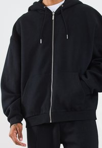 Svart hoodie med glidelås og snorhette, store frontlommer og ribbestrikkede mansjetter. Laget av mykt materiale med en avslappet passform.