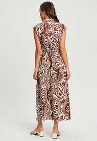 Robe longue sans manches avec un motif tourbillonnant marron et crème, ceinture à la taille et tissu fluide ; possède un col et une longueur maxi.