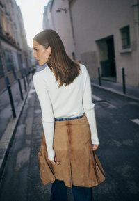 Biała prążkowana bluzka z długim rękawem w połączeniu z brązową zamszową spódnicą kopertową. Spódnica ma plisowany fason, niebieski pas w talii oraz ciemnoniebieskie dżinsy.