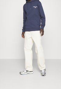 Námořnická mikina s kapucí s logem "TOMMY JEANS", spárovaná s volnými bílými kalhotami a stříbrnými sportovními botami s černými akcenty.