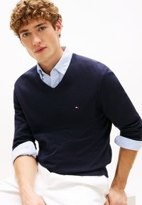 Giovane uomo con capelli ricci che indossa un maglione blu navy a scollo a V sopra una camicia a righe blu con maniche arrotolate e pantaloni bianchi, seduto.