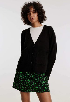 Schwarze Strickjacke mit V-Ausschnitt, ausgestattet mit vier Knöpfen und zwei Fronttaschen, getragen über einem kurzen schwarzen Rock mit grünem Blumenmuster.