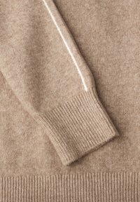 Beige Kaschmirpullover-Detail mit geripptem Bündchen und glatter Textur, ergänzt durch einen kontrastierenden weißen Streifen entlang des Ärmels.