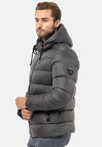 Graue gepolsterte Jacke mit Kapuze, ausgestattet mit einem Reißverschluss vorne und seitlichen Taschen. Auffälliges Logo auf dem linken Arm. Glatte, glänzende Textur.