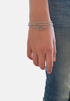 Mano con bracciale avvolgente con perline e ciondolo a forma di loto in argento, posata accanto a una gamba con jeans di denim blu su uno sfondo semplice.