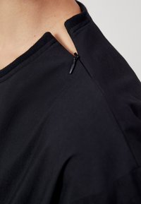 Top in tessuto nero con scollo rotondo, dettaglio con zip piccola su un lato e texture liscia. Design semplice e moderno.