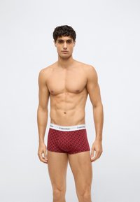 Mann med kort mørkt hår som har på seg rød mønstret Calvin Klein boxershorts, står mot en ensfarget lys grå bakgrunn.