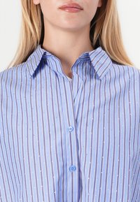 Camicia azzurro chiaro con righe verticali. Il materiale sembra liscio, con sottili accenti metallici e un colletto strutturato.