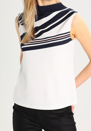 Ærmeløs hvid top med høj krave og diagonale sorte og mørkerøde striber over brystet, båret med sorte bukser.