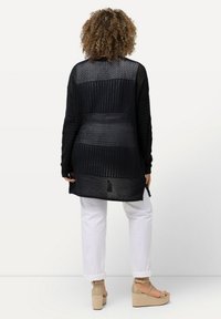 Svart, lätt stickad cardigan med en texturerad, öppen vävdesign, bärs över vita jeans och bruna kilklacksandaler.