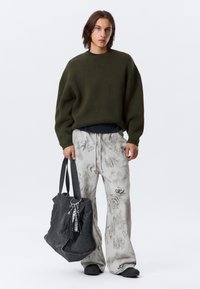 Pull surdimensionné en tricot vert olive, pantalon de survêtement gris clair à motifs, sac fourre-tout noir avec détails en métal, et chaussures noires sans lacets.
