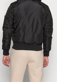 Černá bomber bunda s lesklým, hladkým povrchem, staženými rukávy, žebrovanými manžetami a lemem. Nosit s světle béžovými kalhotami.