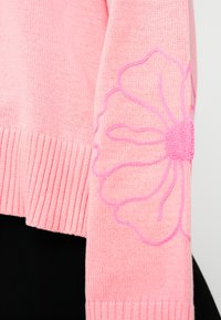 Ροζ πλεκτό πουλόβερ με ριμπ τελείωμα, με μεγάλο κεντητό λουλούδι σε σκούρο ροζ στη αριστερή μανσέτα.