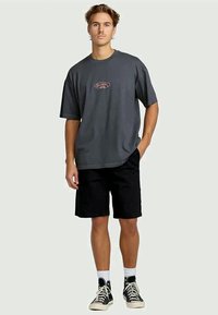 Jonge man in een losse donkergrijze Billabong T-shirt, zwarte shorts, witte sokken en zwarte hoge sneakers, staande met één hand in zijn zak.