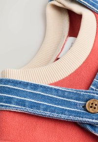Koralfarvet stof med ribbet cremehalsudskæring, der har en blå denimrem med hvide striber og en brun knapdetalje.