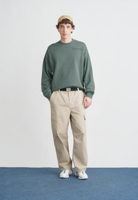 Young man wearing beige cargo pants, green sweatshirt, beige cap, and white sneakers standing against a plain white wall on blue carpet.