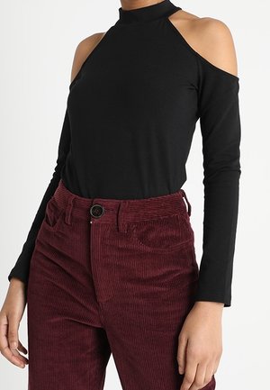 Mustan kylmäsäärisen pitkähihaisen paidan yhdistelmä korkeavyötäröisten burgundinpunaisten, vanutettujen housujen kanssa henkilöllä.