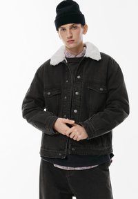 Giacca di jeans nera con colletto in pelliccia sintetica bianca, chiusura con bottoni, due tasche sul petto, maniche lunghe e vestibilità comoda. Indossata sopra un outfit a strati.