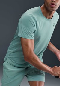 Atletische persoon in een bijpassend lichtgroen T-shirt met korte mouwen en korte broek, die in een bereidheidspositie een tennisracket vasthoudt tegen een donkere achtergrond.
