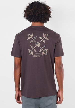 Personne aux cheveux bouclés porte un t-shirt foncé avec un grand motif croisé de fleur et de feuille avec le mot "OXBOW" en dessous dans le dos.