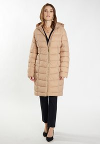 Beige dunjakke med hætte, vertikalt quiltet mønster og frontlynlås med guldfarvet hardware. Båret over sorte bukser og højhælede sko.