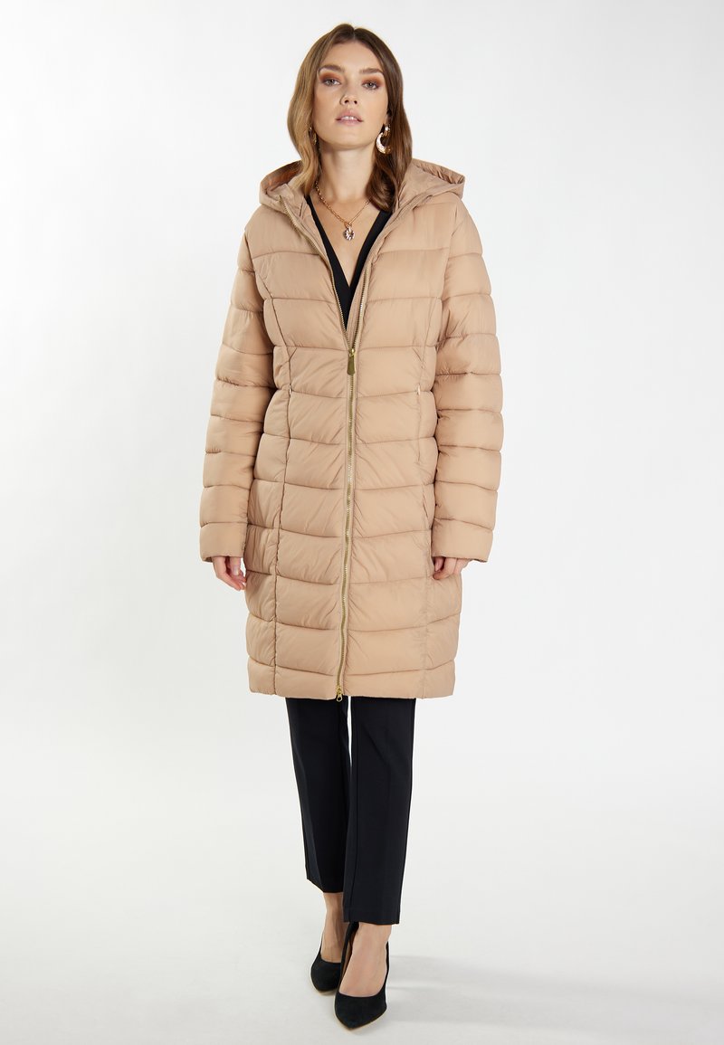 Beige Daunenjacke mit Kapuze, vertikalem Steppmuster und frontalem Reißverschluss mit goldenen Verzierungen. Getragen über schwarzen Hosen und Schuhen mit Absatz.