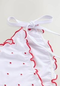 Biele bikiny s červenými volánikmi a malými červenými srdiečkami, s viazaným popruhom na ramene pre nastaviteľné prispôsobenie.