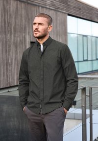 Camisa de botones de patrón oscuro con cuello amplio, mangas largas y tela texturizada. Pantalones grises, manos en los bolsillos, de pie al aire libre.