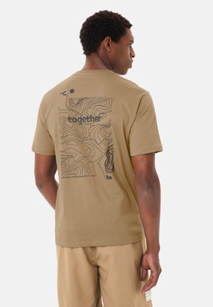 Mann trägt ein beigefarbenes T-Shirt mit schwarzer topografischer Karte und dem Wort "together" auf dem Rücken, kombiniert mit passenden beigefarbenen Hosen.