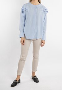Modella che indossa una blusa azzurra chiara con spalle plissettate, pantaloni beige e mocassini neri, in piedi su uno sfondo bianco semplice.
