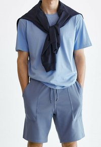 T-shirt bleu clair à col rond, associé à un short de sport bleu clair avec des poches latérales. Un sweat bleu marine foncé noué autour des épaules.