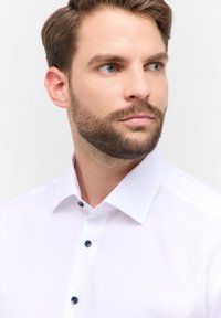 Weißes Hemd mit klassischem Kragen, mit dunklen Knöpfen vorne. Glatte Textur, taillierter Schnitt. Minimalistische Ästhetik.