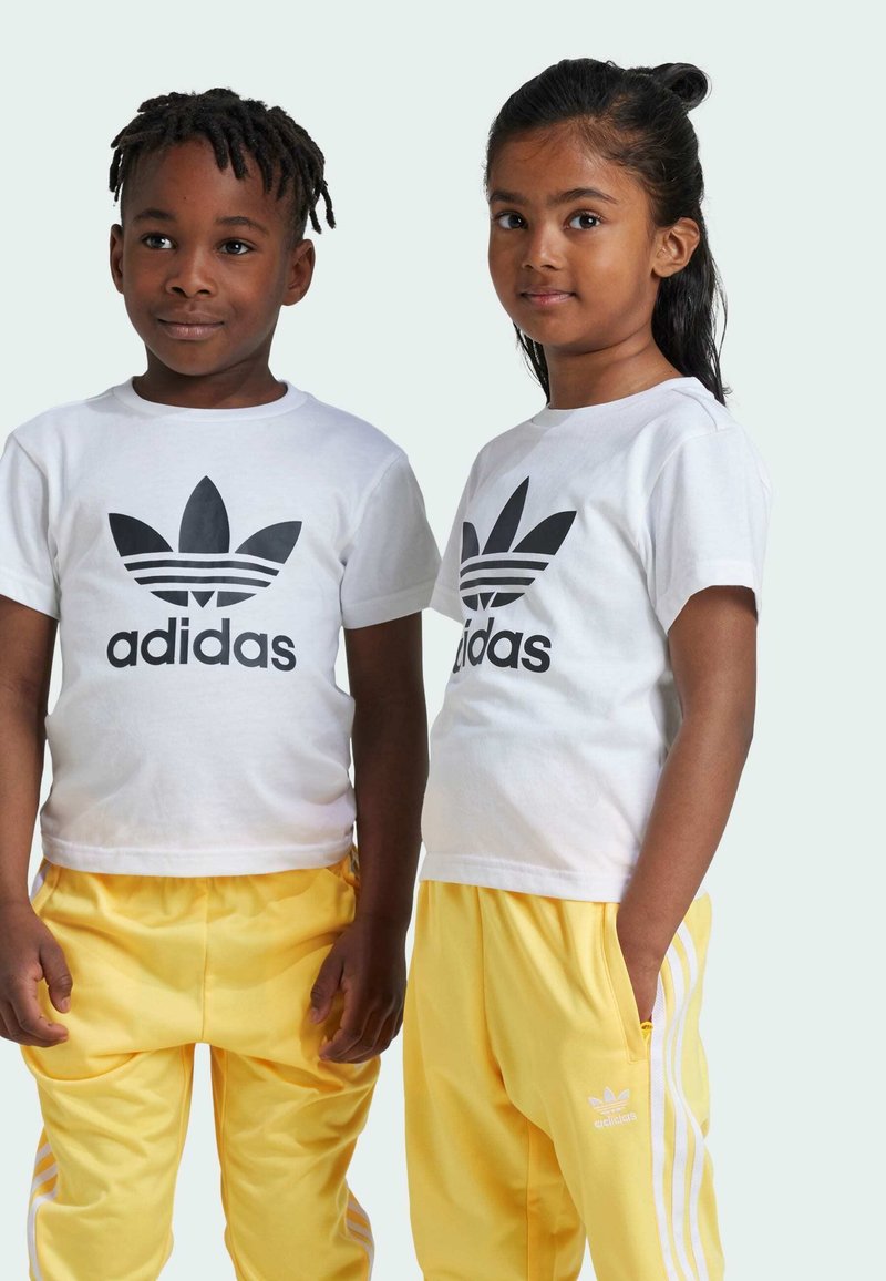 Due bambini indossano t-shirt bianche con il logo Adidas nero e pantaloni da ginnastica gialli con righe bianche sui lati.