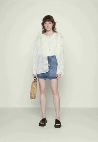 Jovem mulher vestindo uma blusa de renda branca, saia mini de jeans, chinelos pretos e segurando uma bolsa de palha trançada, contra um fundo simples.