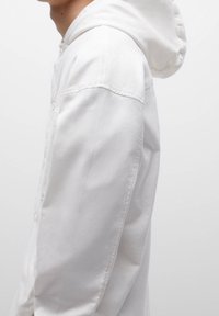 Chaqueta blanca con capucha, con cierre de botones en la parte delantera, textura de tela suave, bolsillos en las costuras laterales y un ajuste clásico. La capucha está ligeramente fruncida en la parte posterior.